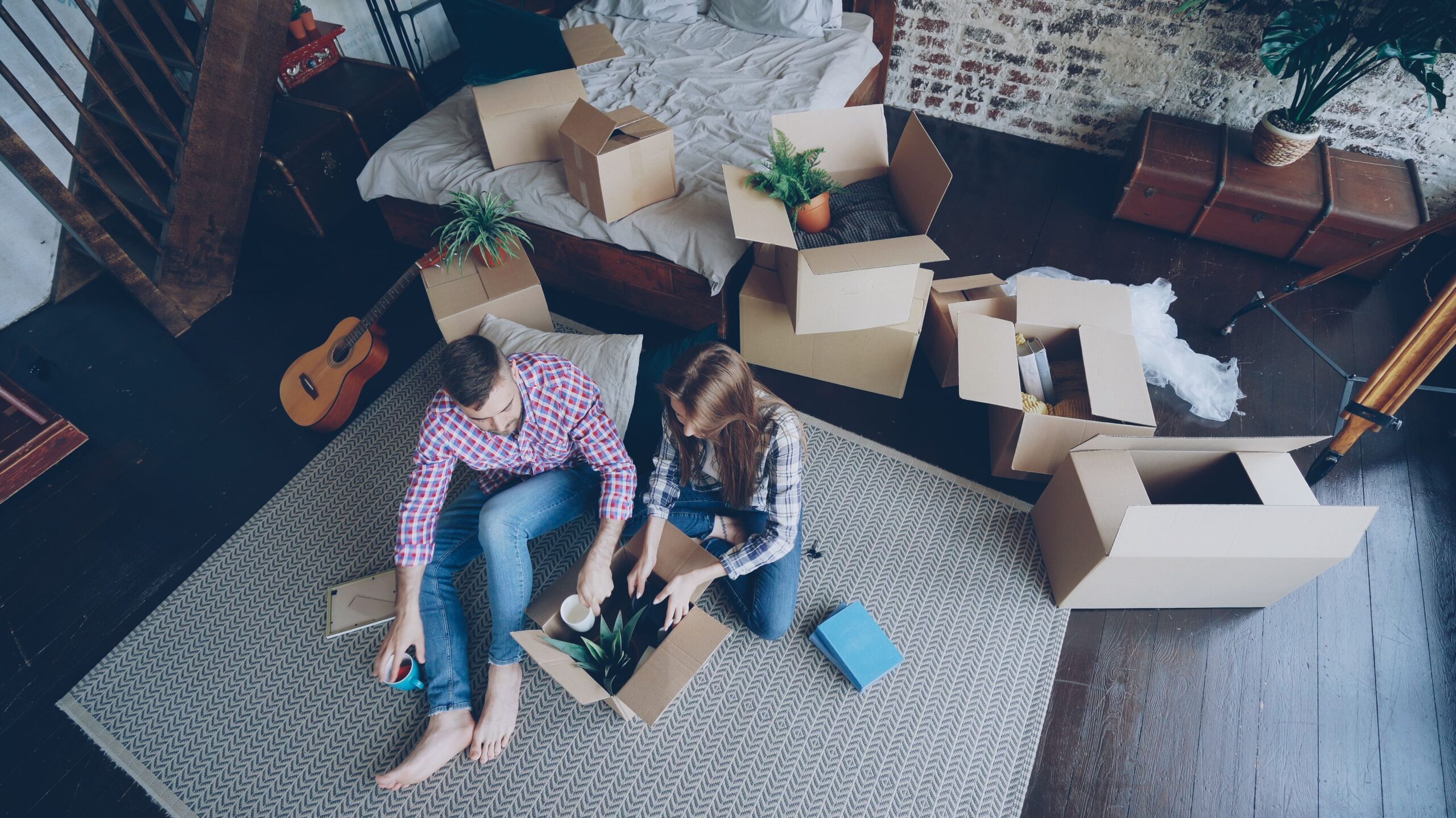 A couple unpacking boxes in a stylish room with plants and a guitar, creating a cozy atmosphere.